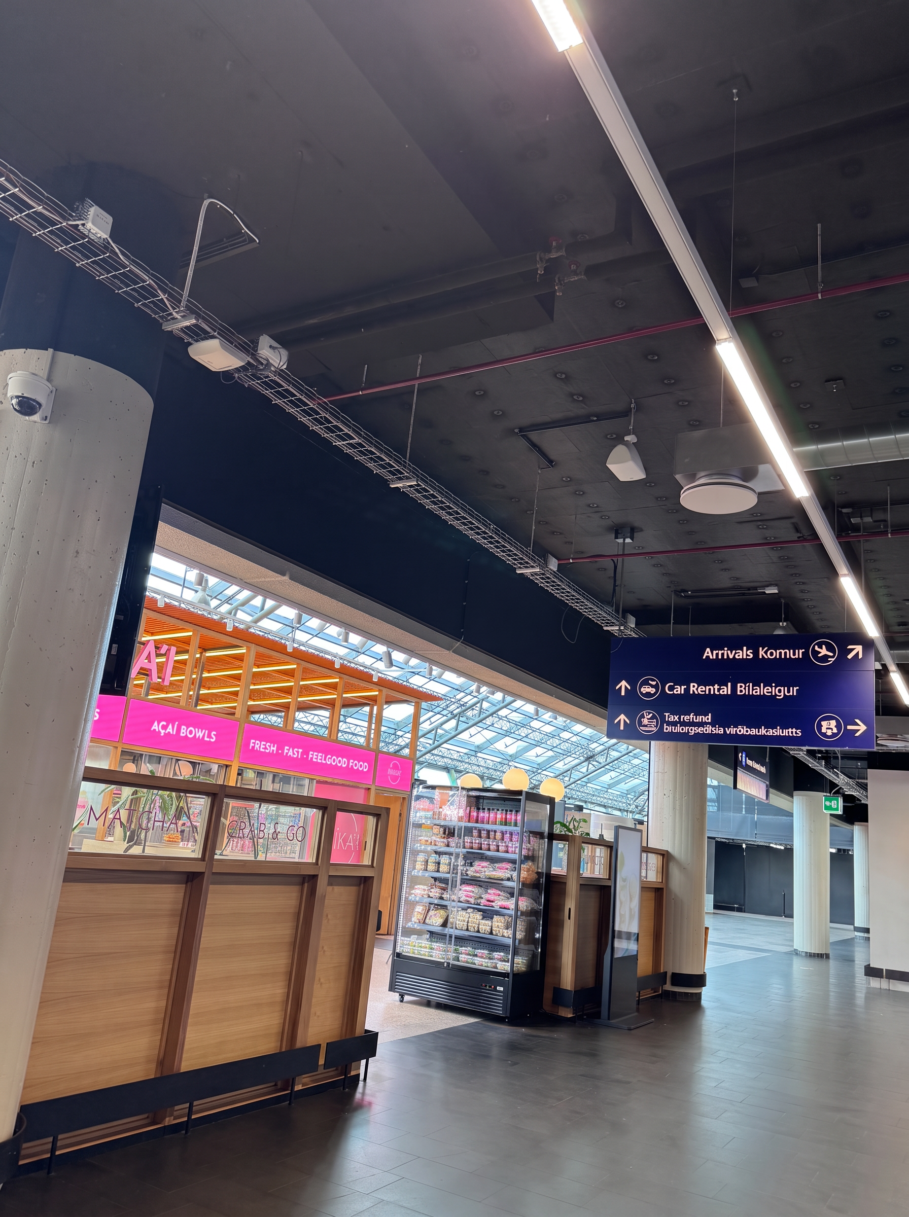 Overhead sign in the Keflavík arrivals hall pointing to Arrivals, Car Rental and Tax refund, beside a coffee and food kiosk