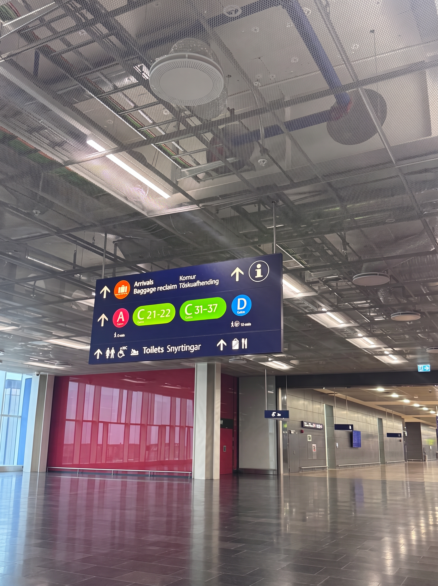 Overhead sign at Keflavík Airport reading Arrivals, Baggage reclaim, Komur, Töskuafhending, with an orange suitcase symbol